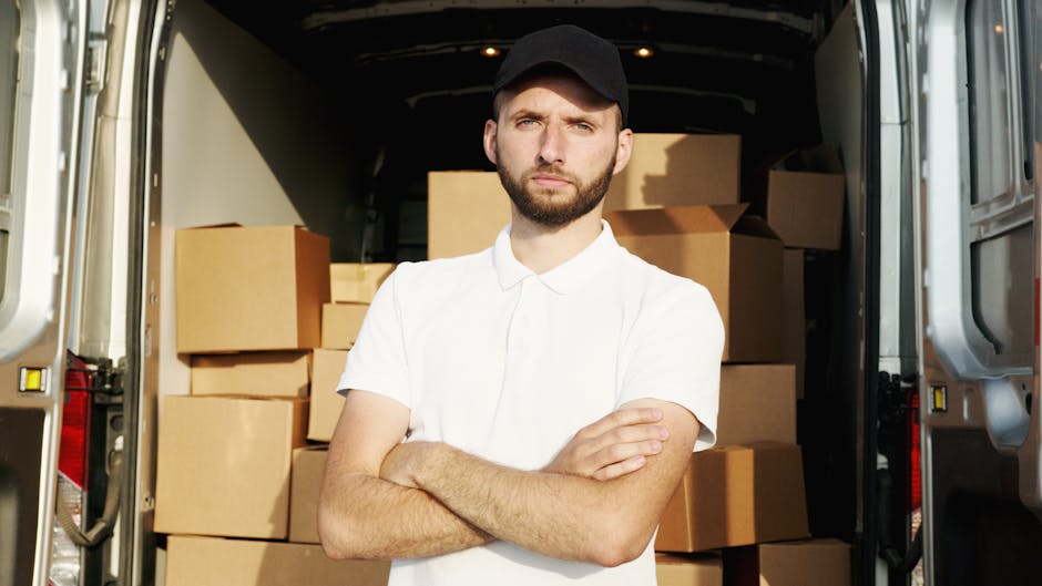A man wearing a white T-shirt and jeans is participating in a home relocation process outside a residential property, standing next to an open moving van with its rear doors swung wide open on the str