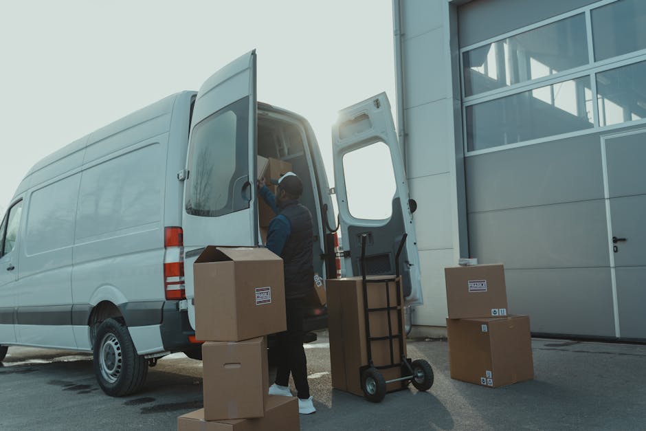 A man dressed in a blue hoodie and navy trousers is loading cardboard boxes into the back of a white van parked on a cobblestone street. The van's rear doors are wide open, revealing an interior lined