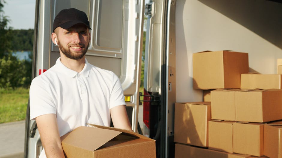 A man wearing a white shirt and glasses is seen unloading cardboard boxes from the back of a moving van parked on a suburban street during sunset. The van's interior is filled with several packed card
