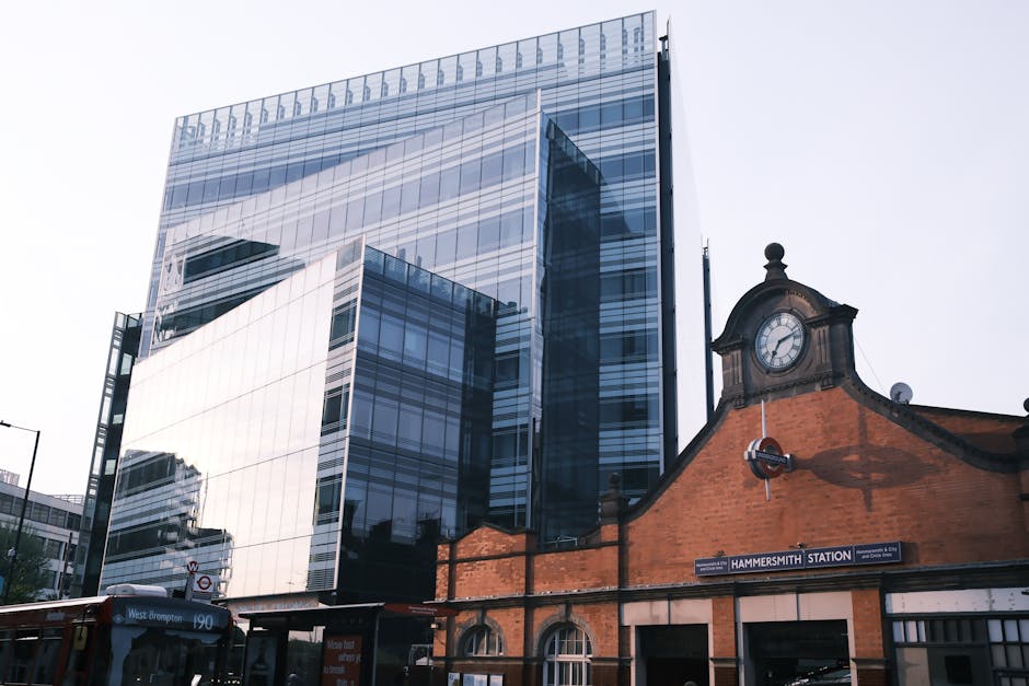 Image showing a modern glass office building with reflective windows towering above a historic brick structure with a clock and a 'Hammersmith Station' sign. In the foreground, part of a bus and stree