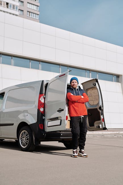 A man wearing a white polo shirt, dark trousers, and a black cap stands inside the open rear of a large moving van, holding a medium-sized cardboard box. The van's interior is filled with numerous oth