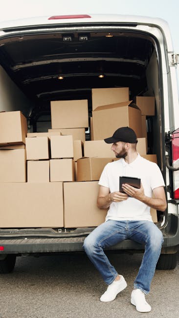 A man wearing a dark padded vest, blue long-sleeve shirt, black trousers, white sneakers, and a white cap stands beside the open rear door of a white moving van parked outside a residential house. The